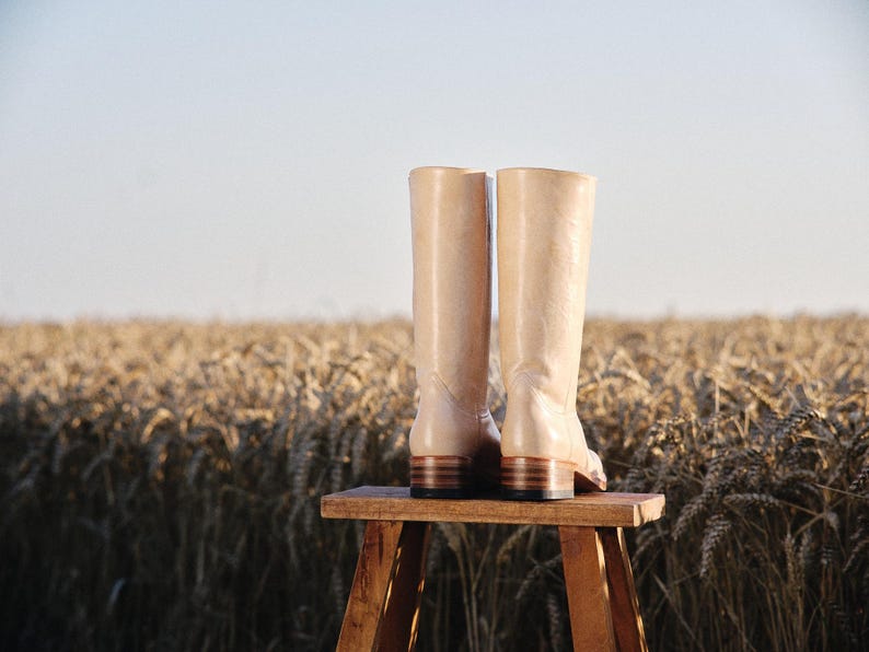 May include: A pair of beige leather boots with a low heel, standing on a small wooden stool. The boots are set against a backdrop of a wheat field under a clear sky, suggesting a rustic or countryside theme.