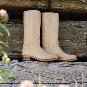 May include: A pair of beige leather cowboy boots. The boots have a classic design with a mid-height shaft, a low heel, and a stitched seam down the front. The boots are set against a rustic wooden background.