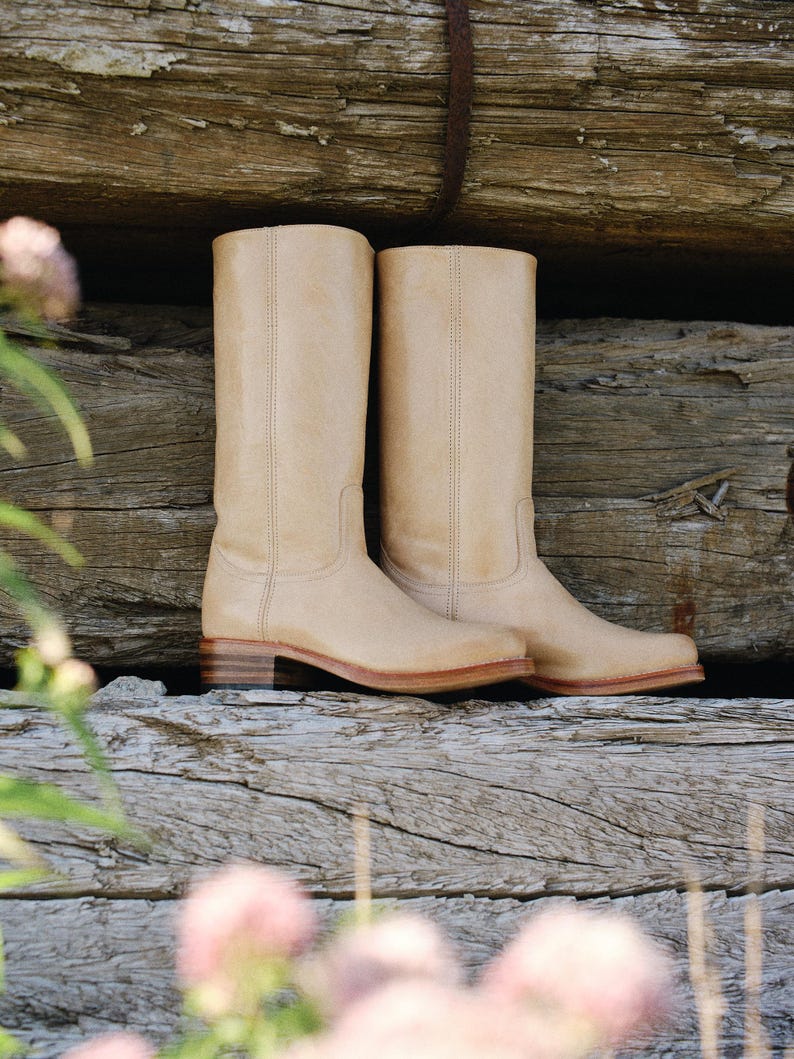 May include: A pair of beige leather cowboy boots. The boots have a classic design with a mid-height shaft, a low heel, and a stitched seam down the front. The boots are set against a rustic wooden background.