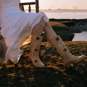 May include: A woman wearing a white dress and floral embroidered cowboy boots sits on a wooden chair overlooking a body of water.