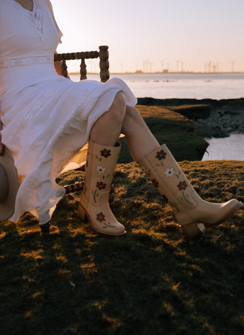 May include: A woman wearing a white dress and floral embroidered cowboy boots sits on a wooden chair overlooking a body of water.