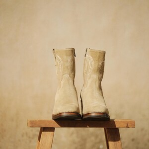May include: A pair of beige suede ankle boots with decorative stitching and zippers. The boots are displayed on a small wooden stool against a neutral background. The boots have a heel height of approximately 1 inch.