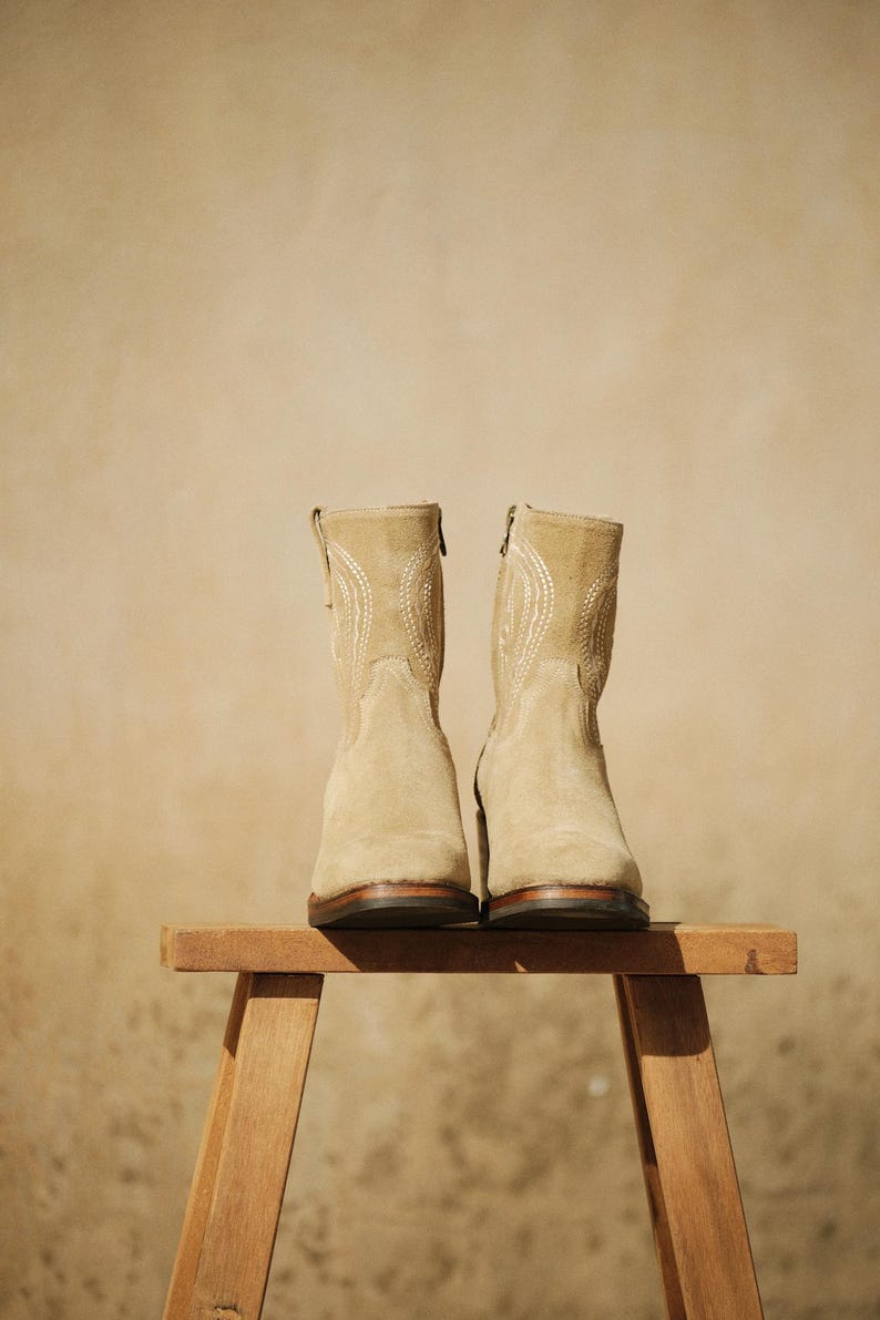 May include: A pair of beige suede ankle boots with decorative stitching and zippers. The boots are displayed on a small wooden stool against a neutral background. The boots have a heel height of approximately 1 inch.