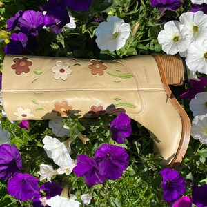 May include: A pair of tan leather cowboy boots with floral embroidery. The boots have a brown heel and a pointed toe. The boots are surrounded by purple and white flowers.