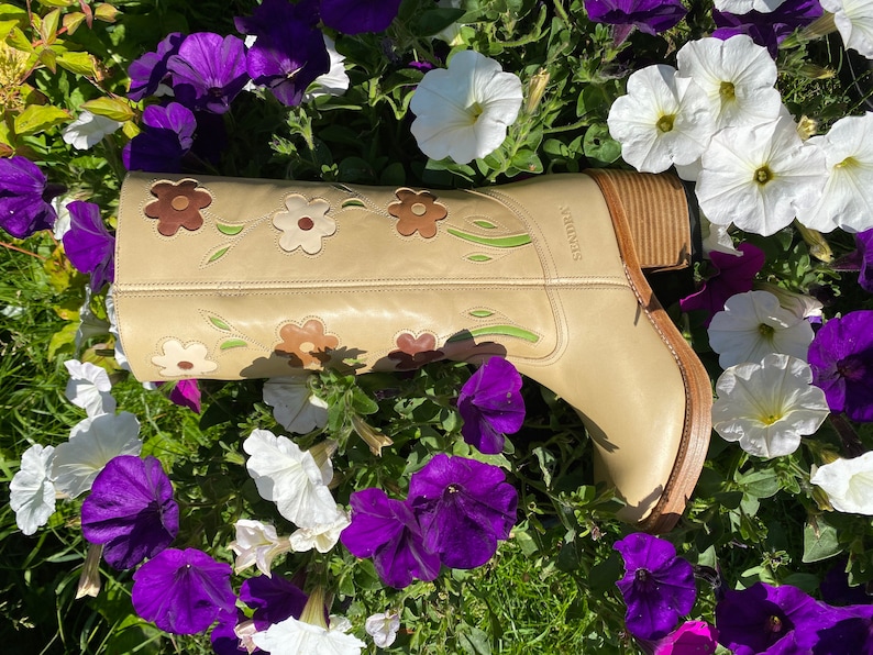 May include: A pair of tan leather cowboy boots with floral embroidery. The boots have a brown heel and a pointed toe. The boots are surrounded by purple and white flowers.