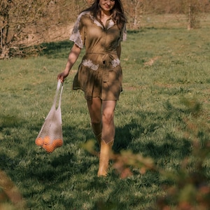 Puede incluir: Una mujer con un vestido marr&oacute;n con adornos de encaje blanco y un sombrero a juego camina por un campo cubierto de hierba. Lleva una bolsa de malla llena de naranjas y botas de vaquero color canela. El vestido tiene una cintura con cintur&oacute;n.