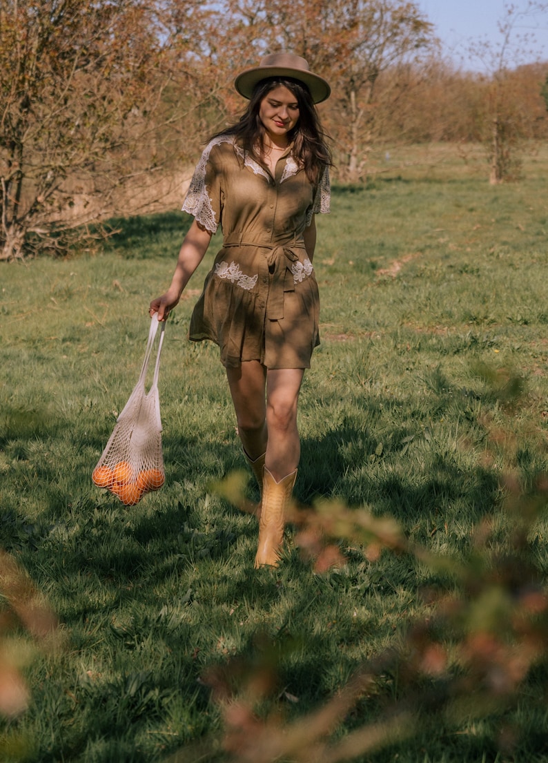 Puede incluir: Una mujer con un vestido marr&oacute;n con adornos de encaje blanco y un sombrero a juego camina por un campo cubierto de hierba. Lleva una bolsa de malla llena de naranjas y botas de vaquero color canela. El vestido tiene una cintura con cintur&oacute;n.