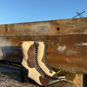 May include: A pair of lace-up ankle boots with a unique cowhide pattern. The boots feature a combination of cream, brown, and black leather. The heels are black, and the toes are pointed. The boots are resting on a wooden surface.