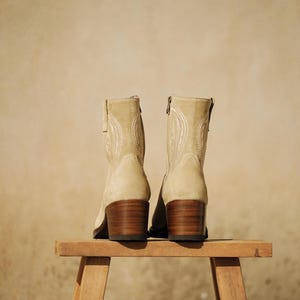 May include: A pair of beige suede ankle boots with decorative stitching and wooden block heels. The boots are displayed on a small wooden stool against a neutral background.