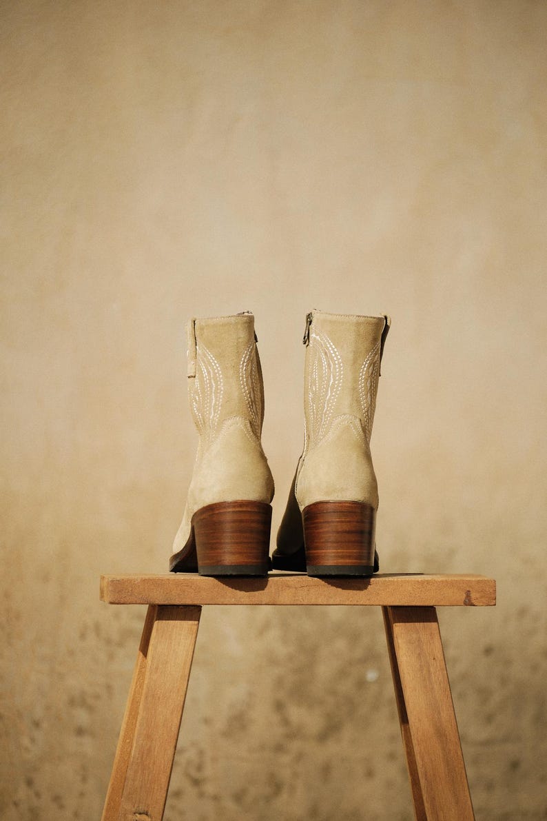 May include: A pair of beige suede ankle boots with decorative stitching and wooden block heels. The boots are displayed on a small wooden stool against a neutral background.