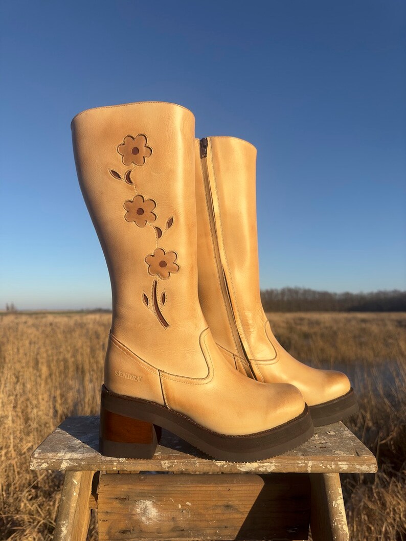 May include: A pair of tan leather knee-high boots with a floral design on the front. The boots have a chunky heel and a platform sole. The brand name "SENDRA" is visible on the side of the boot.