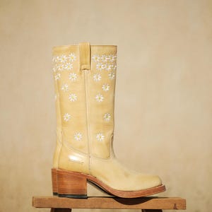 Może przedstawiać: Jasnobrązowy skórzany but kowbojski z kwadratowym noskiem i drewnianym obcasem. But jest ozdobiony białym haftem w stokrotki. But jest prezentowany na małym drewnianym stołku.