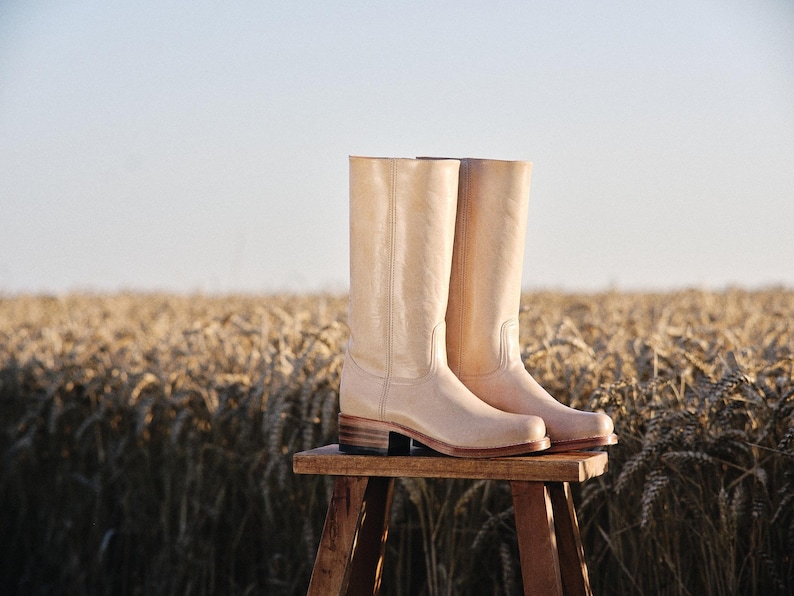 May include: Two light beige leather cowboy boots with stacked heels, placed on a small wooden stool. The boots are tall, reaching the mid-calf, and are set against a backdrop of a wheat field under a clear sky.