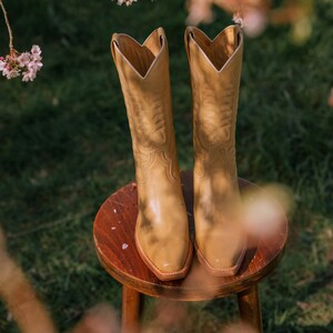Puede incluir: Un par de botas de vaquero color canela con costuras decorativas, apoyadas en un peque&ntilde;o taburete de madera. Las botas tienen una punta puntiaguda y una ca&ntilde;a alta. El fondo presenta hierba verde y flores rosas borrosas.