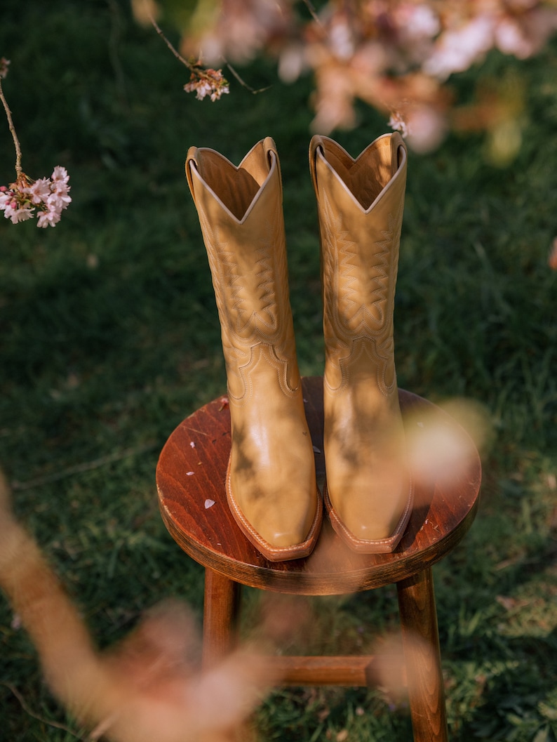 Puede incluir: Un par de botas de vaquero color canela con costuras decorativas, apoyadas en un peque&ntilde;o taburete de madera. Las botas tienen una punta puntiaguda y una ca&ntilde;a alta. El fondo presenta hierba verde y flores rosas borrosas.
