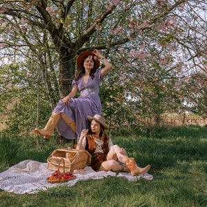 May include: Two people are enjoying a picnic under a blossoming tree. One person wears a purple dress and brown boots, the other wears a brown vest and cowboy boots. A picnic basket and blanket are on the grass.