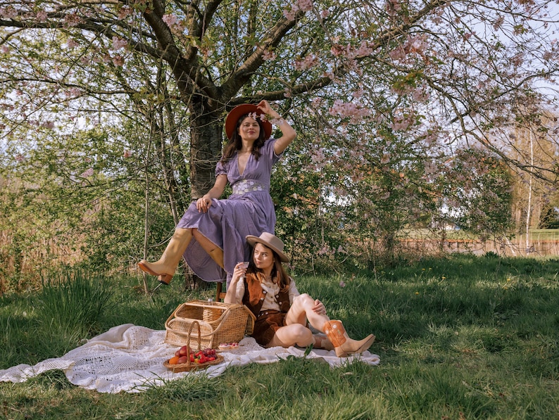 May include: Two people are enjoying a picnic under a blossoming tree. One person wears a purple dress and brown boots, the other wears a brown vest and cowboy boots. A picnic basket and blanket are on the grass.
