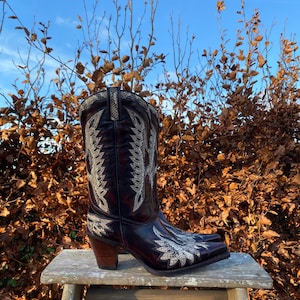 Op de afbeelding: Een paar bruine leren cowboylaarzen met witte borduursels. De laarzen hebben een spitse neus en een hoge hak. De laarzen staan op een houten kruk tegen een achtergrond van bruine bladeren.