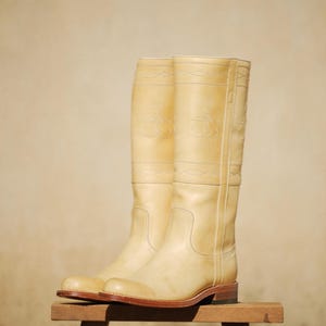 May include: A pair of tall, cream-colored leather riding boots. The boots have decorative stitching and embossed designs near the top. They are displayed on a small wooden stool against a neutral background.