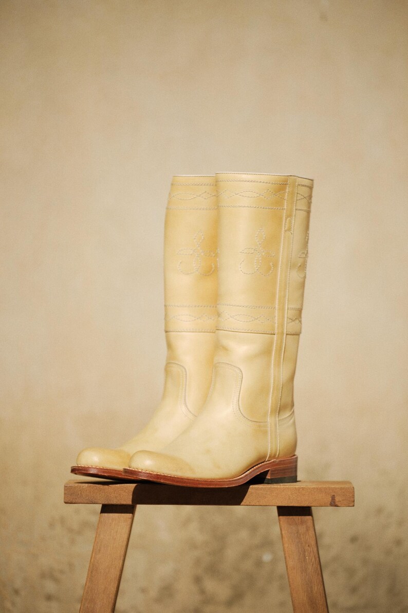May include: A pair of tall, cream-colored leather riding boots. The boots have decorative stitching and embossed designs near the top. They are displayed on a small wooden stool against a neutral background.