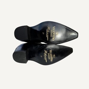 May include: Black leather boots with pointed toes and block heels. The soles display the Valentino Garavani logo, "Hecho a mano MADE IN SPAIN" and size 37. The boots are set against a neutral background.