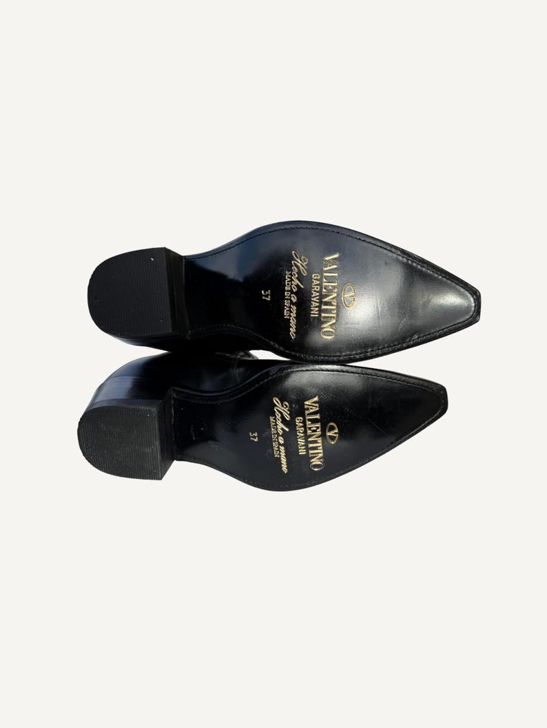May include: Black leather boots with pointed toes and block heels. The soles display the Valentino Garavani logo, "Hecho a mano MADE IN SPAIN" and size 37. The boots are set against a neutral background.