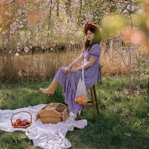 May include: A picnic scene with a woman in a purple dress and brown hat sitting on a wooden stool. A wicker picnic basket, a basket of fruit, and a white blanket are on the grass. A net bag with oranges hangs from the stool.