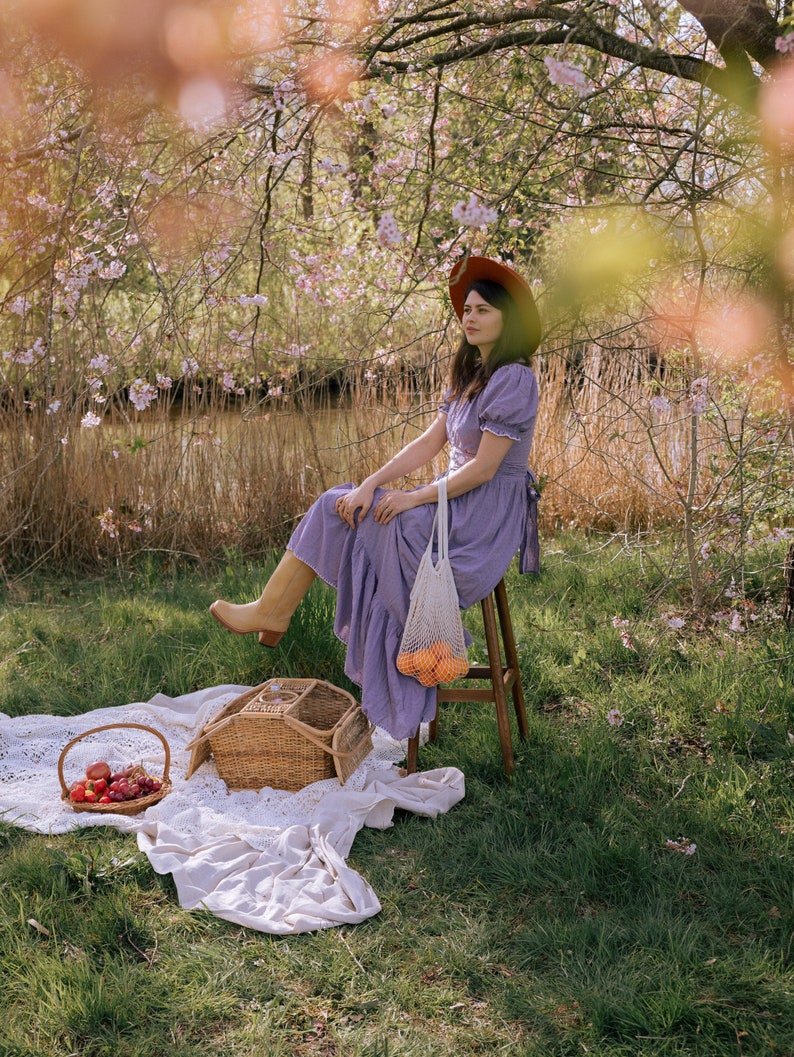 May include: A picnic scene with a woman in a purple dress and brown hat sitting on a wooden stool. A wicker picnic basket, a basket of fruit, and a white blanket are on the grass. A net bag with oranges hangs from the stool.
