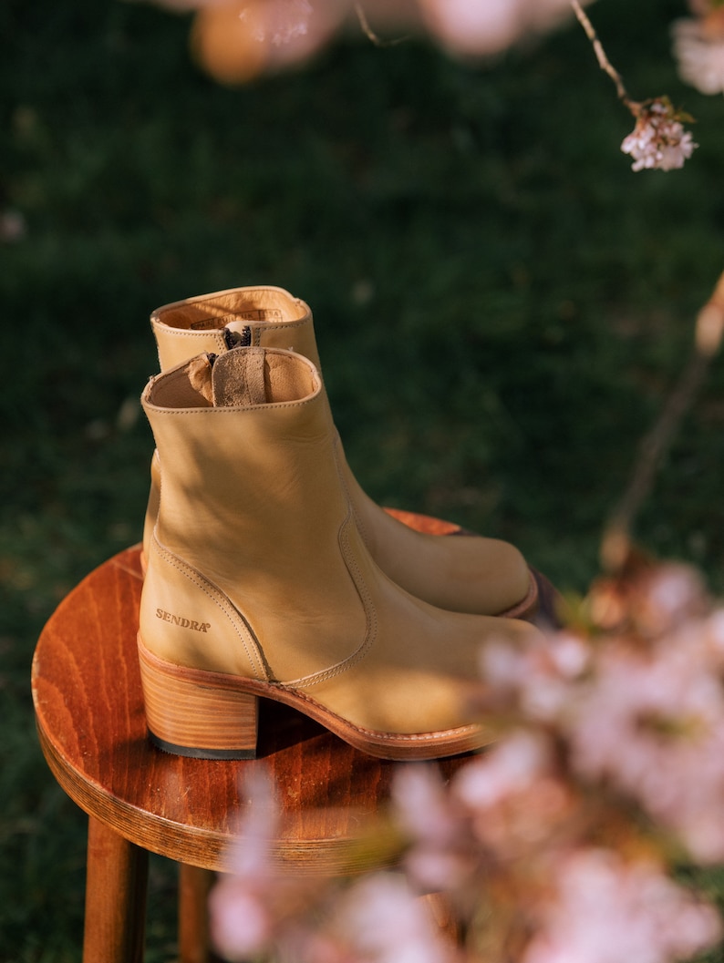 May include: Tan leather ankle boots with a block heel and a side zip. The boots are sitting on a wooden stool with a light brown finish. The brand name "SENDRA" is visible on the side of the boot.