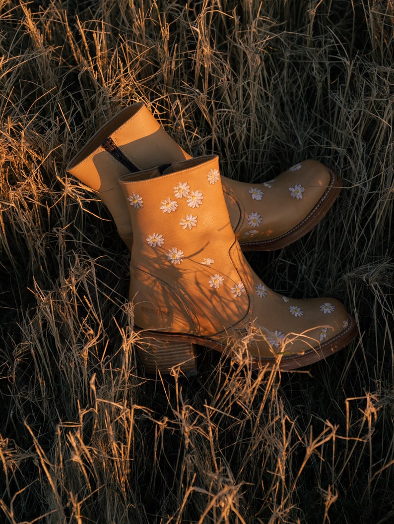 May include: A pair of brown leather ankle boots with white daisy embroidery. The boots are set against a backdrop of dry grass.
