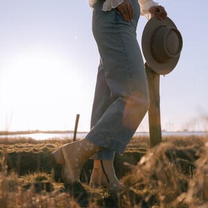 May include: A person wearing a white blouse and blue denim overalls stands in a field of tall grass. They are holding a brown felt hat in their right hand. The person is wearing brown boots with white floral embroidery.