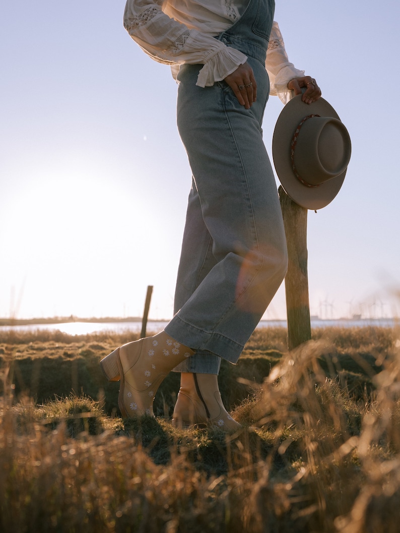 May include: A person wearing a white blouse and blue denim overalls stands in a field of tall grass. They are holding a brown felt hat in their right hand. The person is wearing brown boots with white floral embroidery.