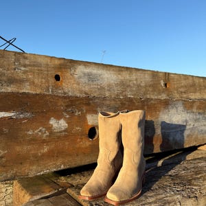 Puede incluir: Un par de botas de vaquero de ante beige con punta cuadrada y tac&oacute;n apilado. Las botas se colocan sobre un fondo de vigas de madera desgastadas y un cielo azul claro, mostrando una est&eacute;tica r&uacute;stica.