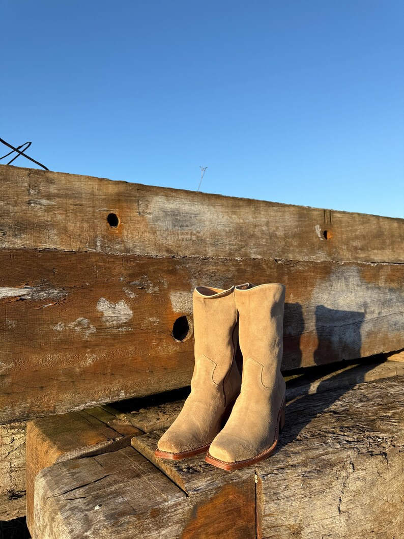 Puede incluir: Un par de botas de vaquero de ante beige con punta cuadrada y tac&oacute;n apilado. Las botas se colocan sobre un fondo de vigas de madera desgastadas y un cielo azul claro, mostrando una est&eacute;tica r&uacute;stica.