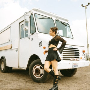 May include: A woman wearing a black crop top and a black mini skirt stands in front of a white delivery truck. She is wearing black boots with floral embroidery. The truck has a California license plate that reads "4THW200".