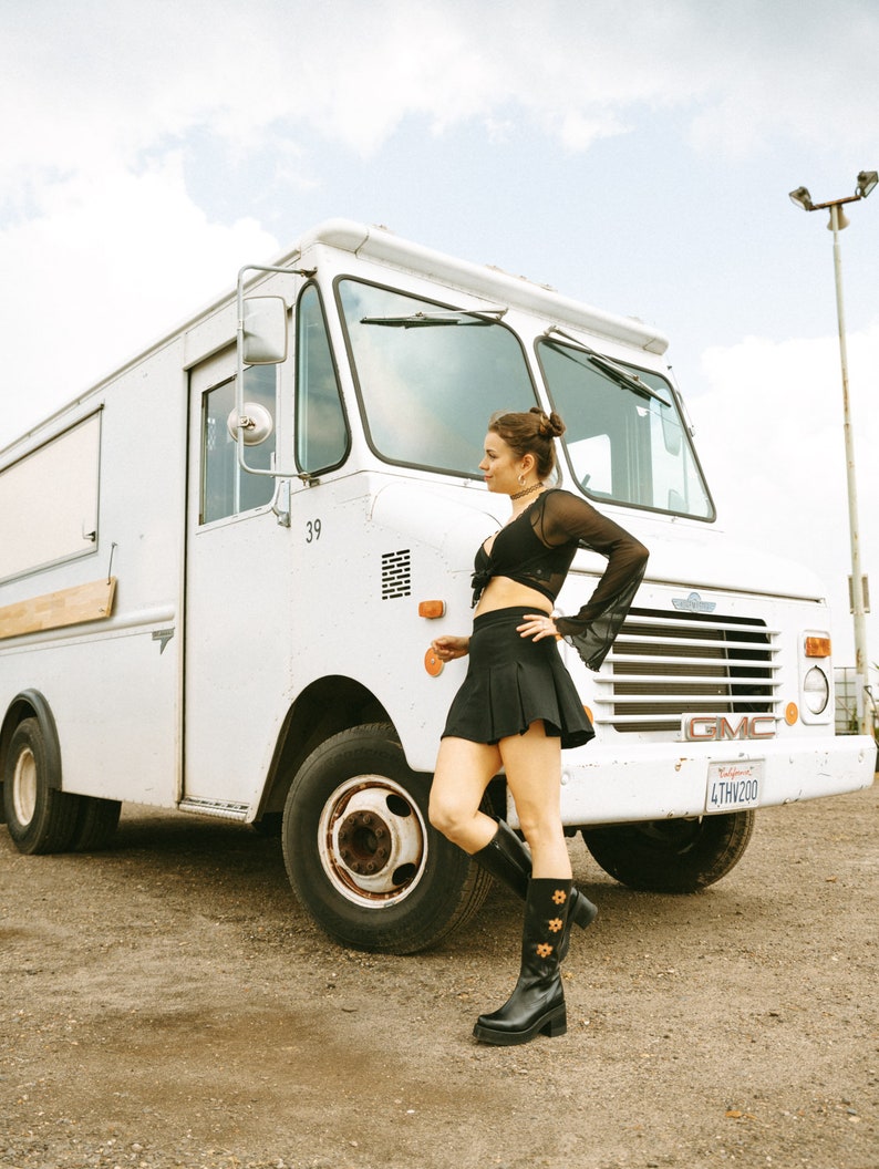 May include: A woman wearing a black crop top and a black mini skirt stands in front of a white delivery truck. She is wearing black boots with floral embroidery. The truck has a California license plate that reads "4THW200".