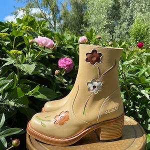 Może przedstawiać: Beżowe skórzane botki z grubym obcasem i kwiatowymi wycięciami. Botki mają brązową skórzaną podeszwę i są ozdobione zielonymi i pomarańczowymi kwiatami.