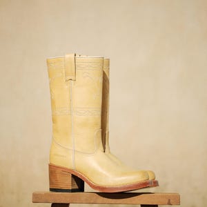 Puede incluir: Botas de vaquero de color amarillo claro con costuras decorativas. Las botas tienen punta cuadrada y tac&oacute;n de madera, expuestas sobre un peque&ntilde;o taburete de madera. Las botas miden unos 30 cm de alto.