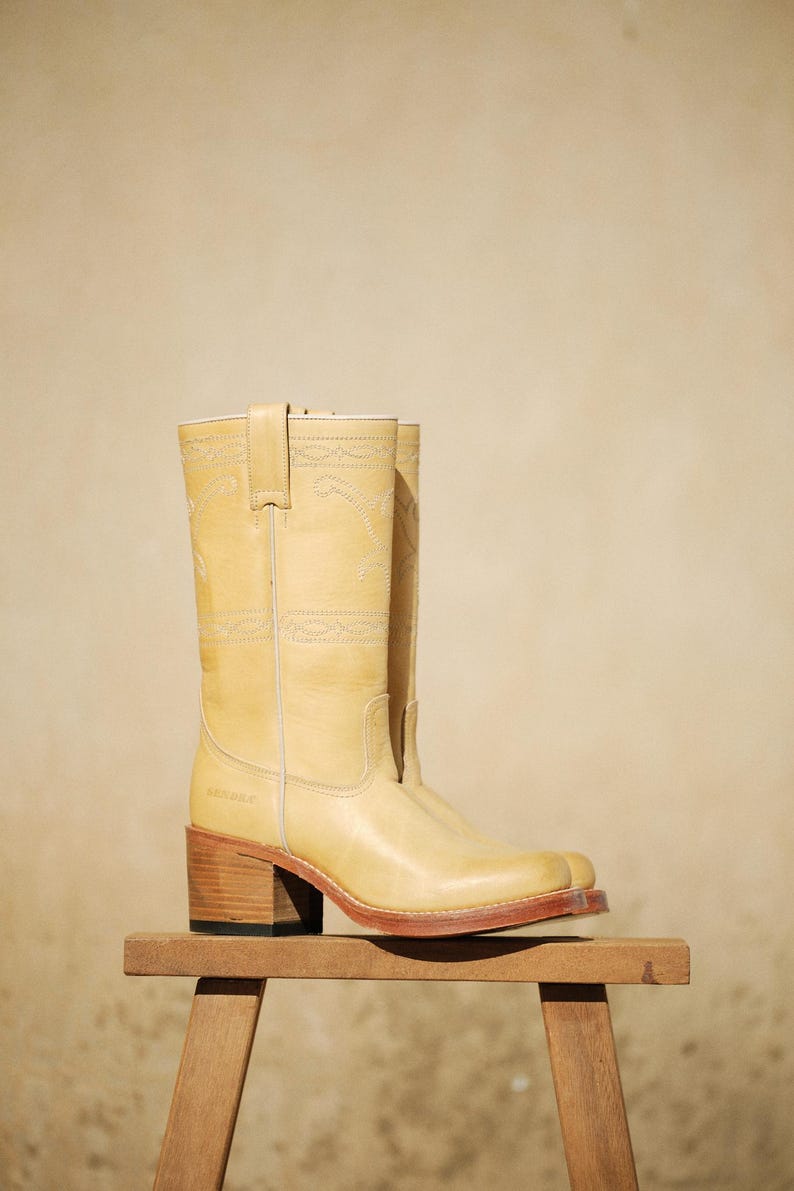 Puede incluir: Botas de vaquero de color amarillo claro con costuras decorativas. Las botas tienen punta cuadrada y tac&oacute;n de madera, expuestas sobre un peque&ntilde;o taburete de madera. Las botas miden unos 30 cm de alto.