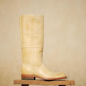 May include: A tall, light beige leather boot with decorative stitching and a low heel. The boot is displayed on a small wooden stool against a neutral background. The boot's design includes stitched floral patterns.