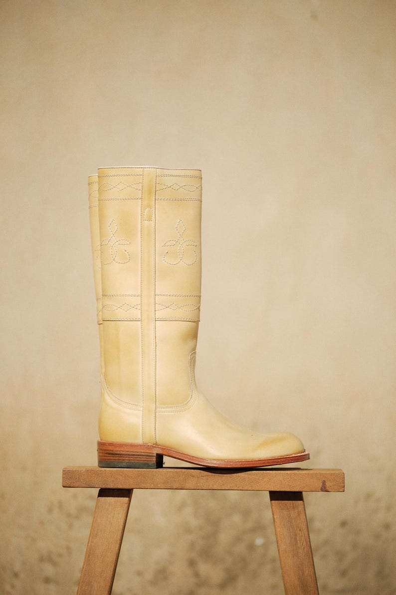 May include: A tall, light beige leather boot with decorative stitching and a low heel. The boot is displayed on a small wooden stool against a neutral background. The boot's design includes stitched floral patterns.