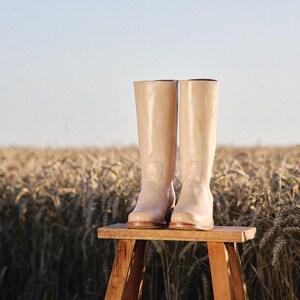 May include: A pair of tall, light beige boots with a glossy finish, standing on a small wooden stool. The boots are the focal point, set against a backdrop of a wheat field under a clear sky. The boots appear to be made of a smooth material.