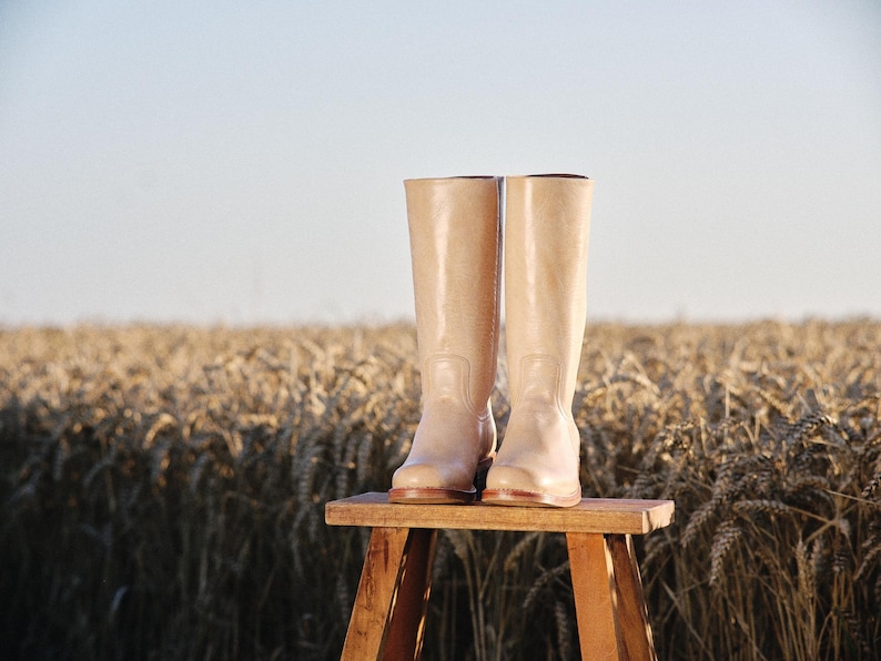 May include: A pair of tall, light beige boots with a glossy finish, standing on a small wooden stool. The boots are the focal point, set against a backdrop of a wheat field under a clear sky. The boots appear to be made of a smooth material.