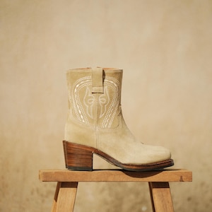 May include: A beige suede ankle boot with a decorative white stitched design. The boot has a stacked wooden heel and is displayed on a small wooden stool. The boot's design is reminiscent of a cowboy boot.