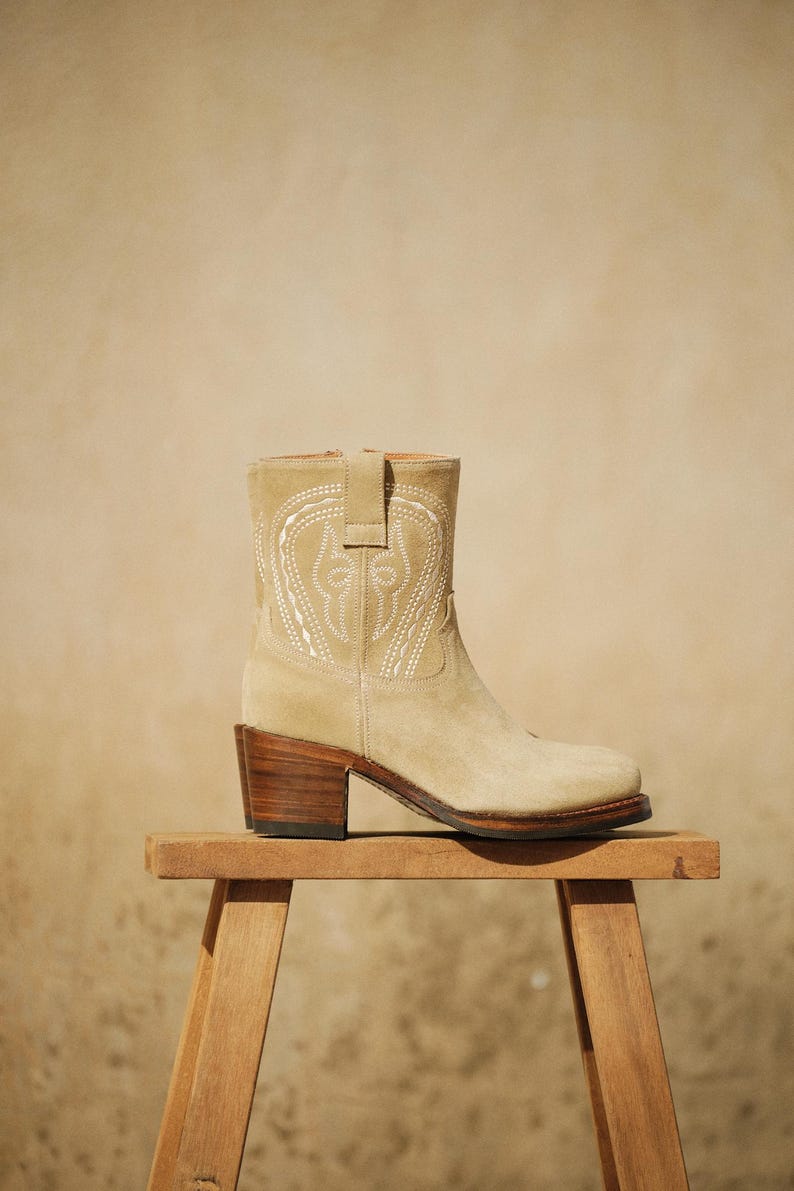 May include: A beige suede ankle boot with a decorative white stitched design. The boot has a stacked wooden heel and is displayed on a small wooden stool. The boot's design is reminiscent of a cowboy boot.