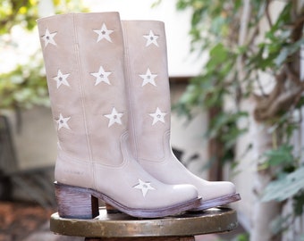 May include: A pair of beige leather cowboy boots with white star cutouts. The boots have a block heel and are sitting on a wooden stool.