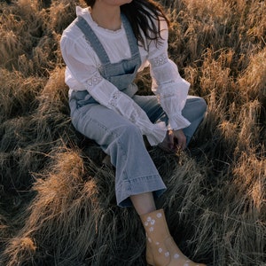 May include: A woman wearing a white lace blouse, blue denim overalls, a gray fedora, and brown boots with white floral embroidery sits in a field of dry grass.