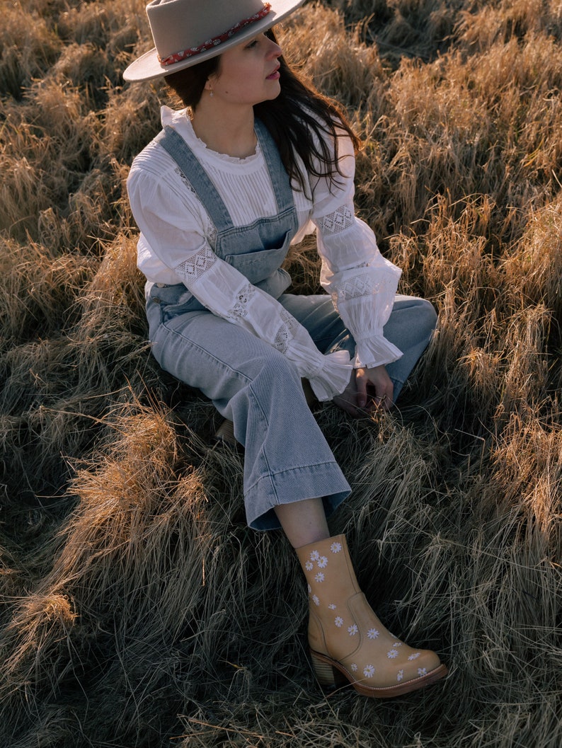 May include: A woman wearing a white lace blouse, blue denim overalls, a gray fedora, and brown boots with white floral embroidery sits in a field of dry grass.
