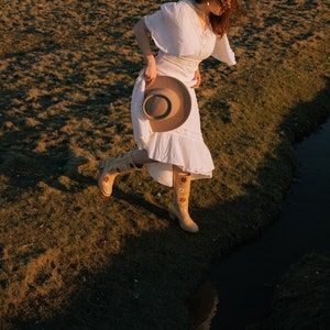 May include: A woman in a white dress and floral boots walks through a grassy field with a brown hat in her hand.