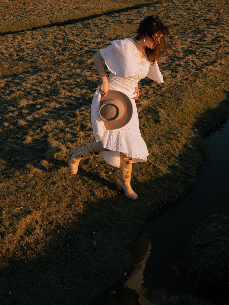 May include: A woman in a white dress and floral boots walks through a grassy field with a brown hat in her hand.